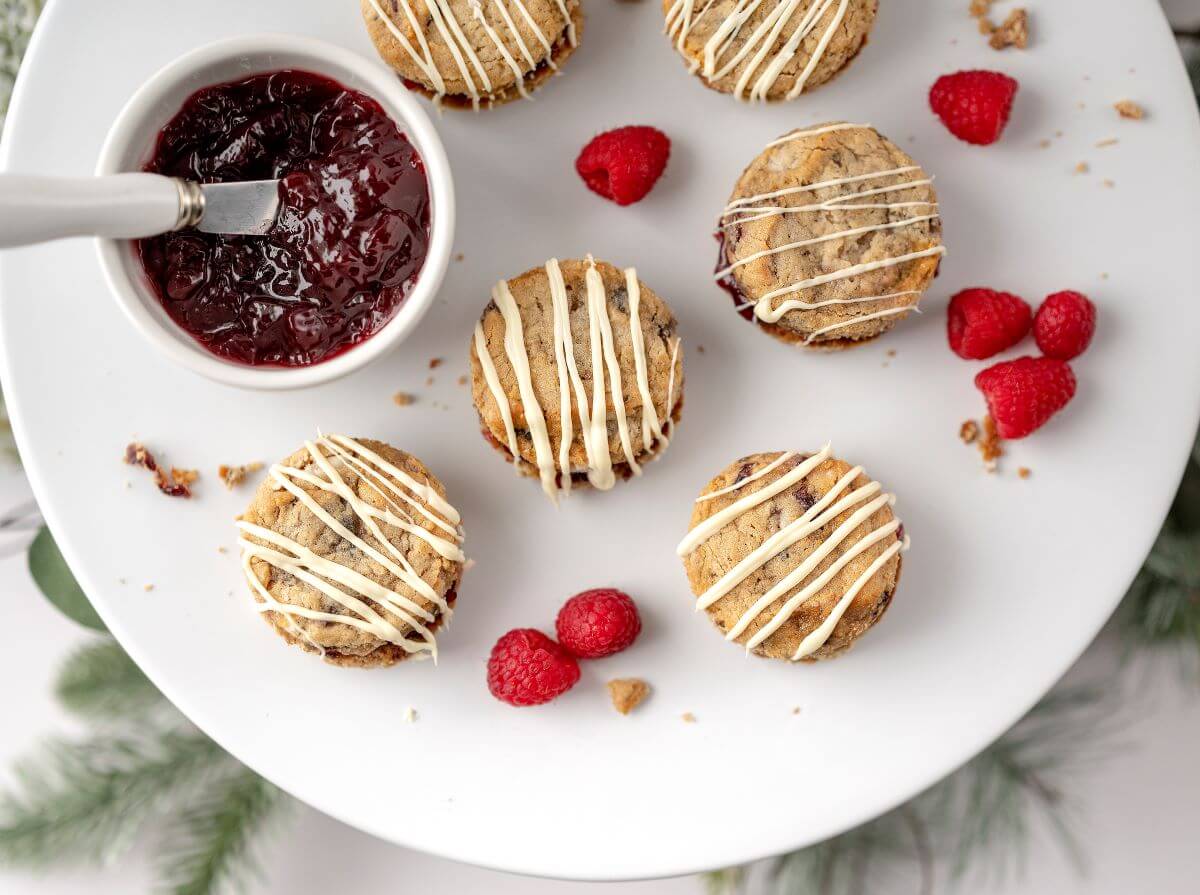 Oatmeal White Chocolate Raspberry Cookie Sandwiches on plate next to bowl of jam and surrounded by fresh raspberries.