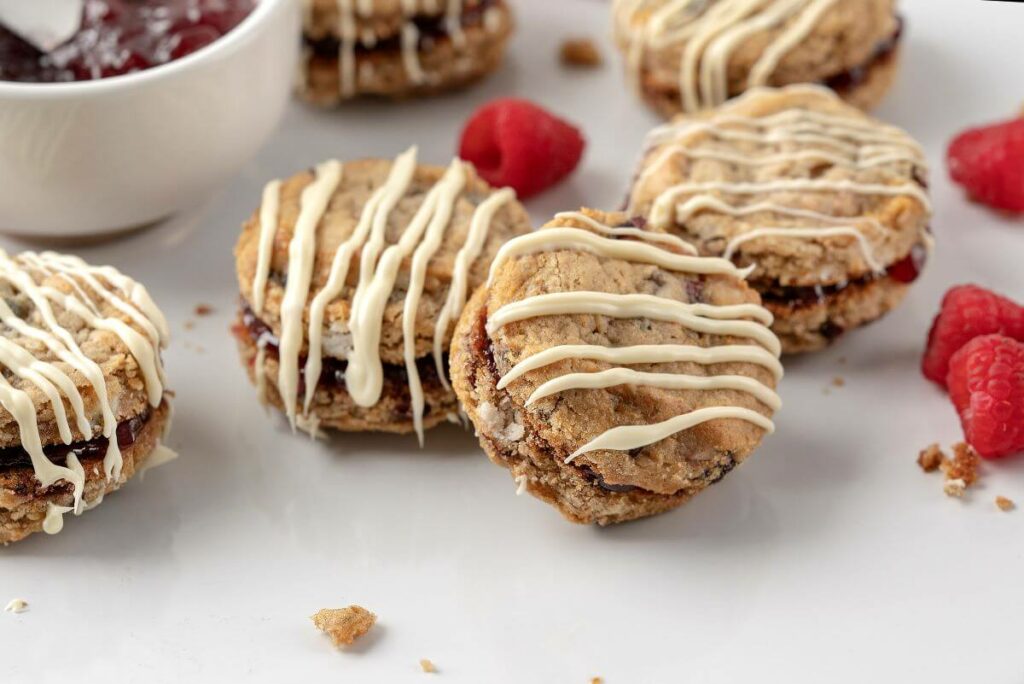 Close up of Oatmeal White Chocolate Raspberry Cookie Sandwiches on plate next to bowl of jam and surrounded by fresh raspberries.