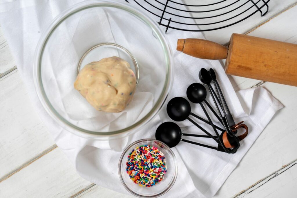Ingredients to make Confetti Donut Cookies - cookie dough in a bowl, a rolling pin, measuring spoons, and a bowl of sprinkles.