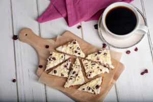 Cranberry Bliss Bars on cutting board next to a cup of coffee and purple cloth napkin. Craisins are scattered around on a white wooden surface.