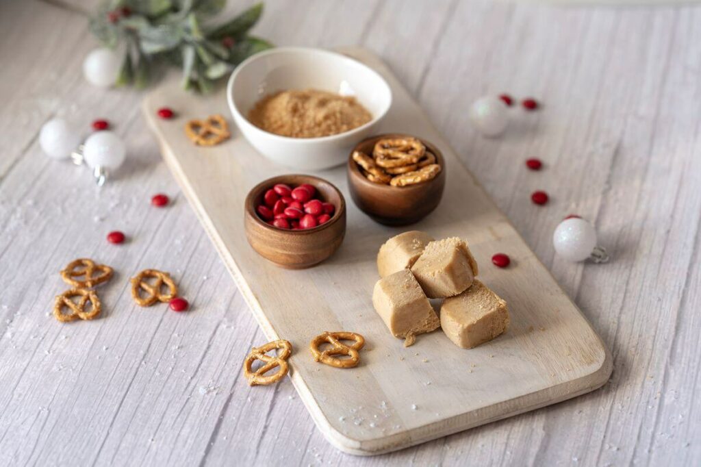 Ingredients for the Rudolph Peanut Butter Cookies - sugar, pretzels, red chocolate candies, and peanut butter cookie dough cubes on cutting board.
