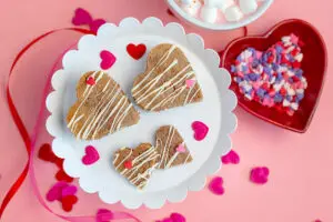 Piñata Valentine's Day Cookies on a white stand next to a bowl of sprinkles and a cup of pink hot chocolate. Valentine's Day decorations are scattered around the stand.