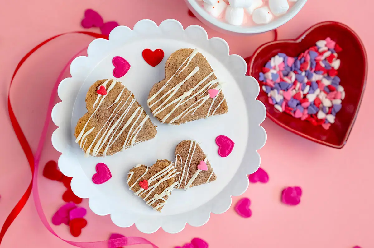 Piñata Valentine's Day Cookies on a white stand next to a bowl of sprinkles and a cup of pink hot chocolate. Valentine's Day decorations are scattered around the stand.
