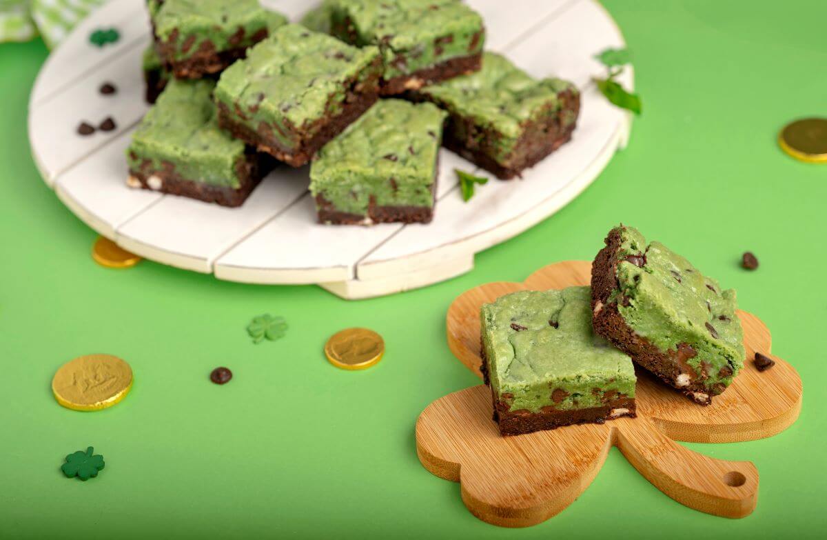 Triple Chocolate Mint Brookies - several on a white wood serving platter in the background of the image. In the foreground, there's two brookies on a shamrock-shaped board. There's chocolate gold coins and shamrock confetti scattered on the green background.