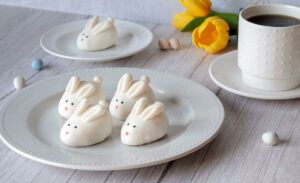 Four Easter Bunny Cookie Truffles on a plate near the forefront of the image. In the background, there's another cookie truffle on a smaller plate near a cup of coffee and yellow tulips.