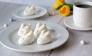 Four Easter Bunny Cookie Truffles on a plate near the forefront of the image. In the background, there's another cookie truffle on a smaller plate near a cup of coffee and yellow tulips.