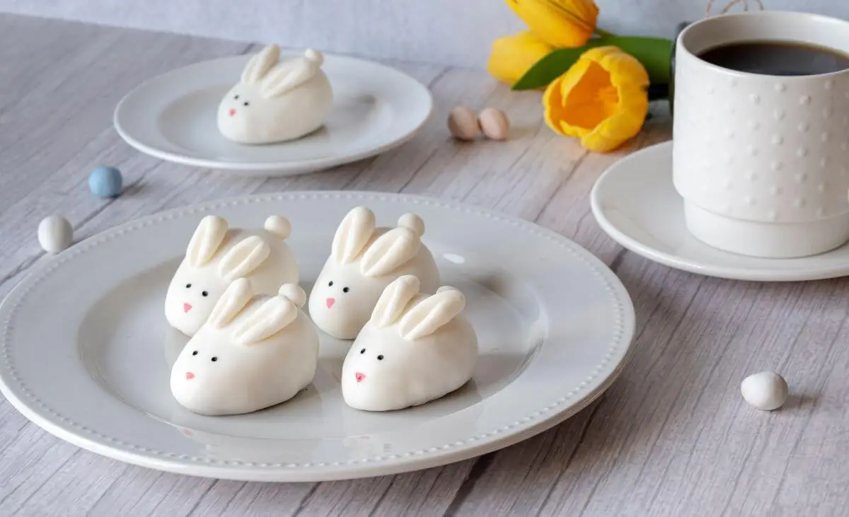 Four Easter Bunny Cookie Truffles on a plate near the forefront of the image. In the background, there's another cookie truffle on a smaller plate near a cup of coffee and yellow tulips.