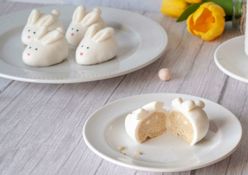 In the background, there are four Easter Bunny Cookie Truffles on a plate. In the foreground, there's another cookie truffle on a smaller plate; this truffle has been cut in half to show the inside.