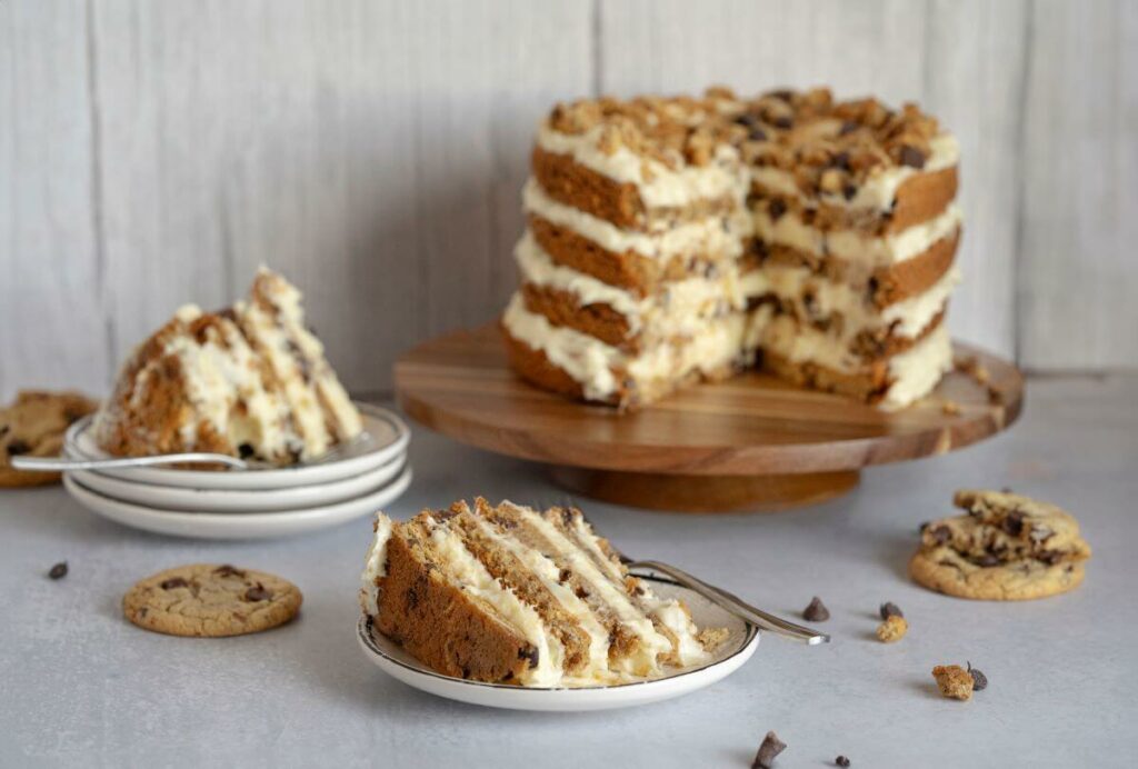 Chocolate Chip Cookie Cake - slices on plates in the foreground of the image with the full layer cake in the back. All surrounded by chocolate chip cookies.