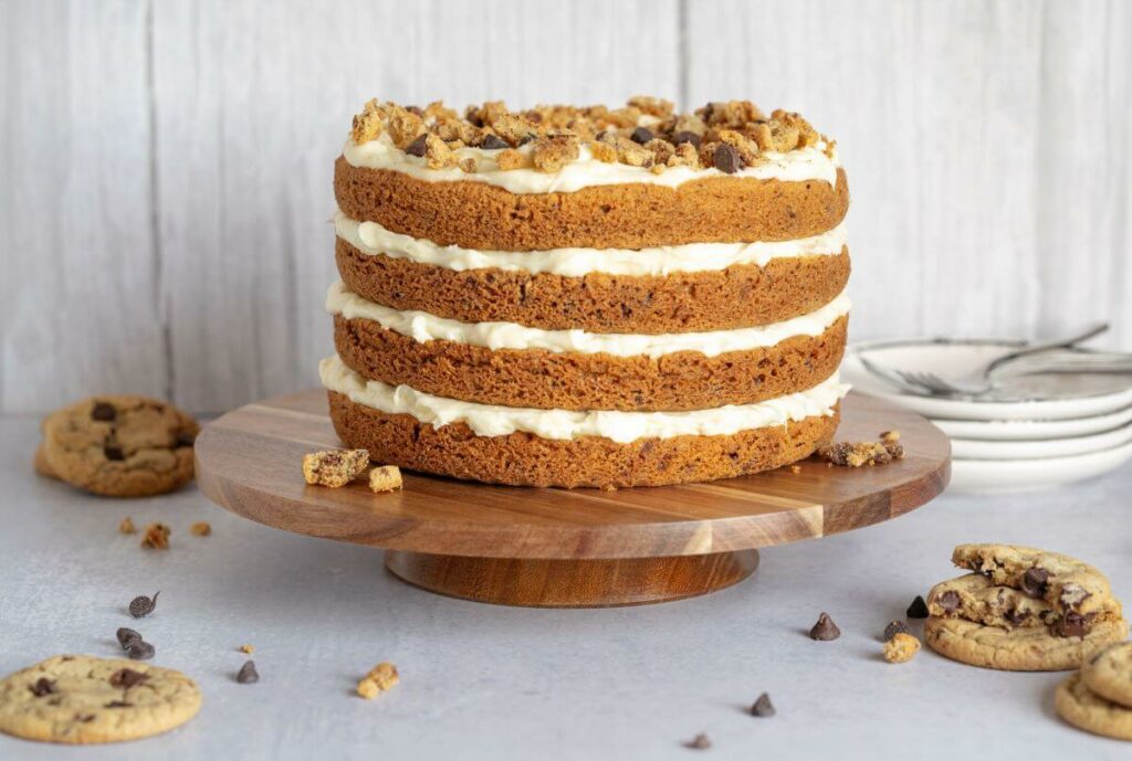 Chocolate Chip Cookie Cake on a cake stand surrounded by chocolate chip cookies.
