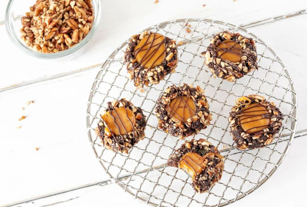 5 full Turtle Thumbprint Cookies on a wire rack and one cookie with a bite taken out of it. A bowl of chopped pecans is in the background.