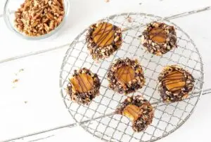 5 full Turtle Thumbprint Cookies on a wire rack and one cookie with a bite taken out of it. A bowl of chopped pecans is in the background.