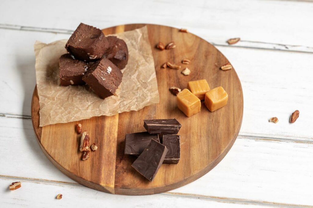 A wooden cutting board with some of the ingredients used to make Turtle Thumbprint Cookies including Triple Chocolate Cookie Dough cubes, caramel bits, semi-sweet chocolate, and pecans.