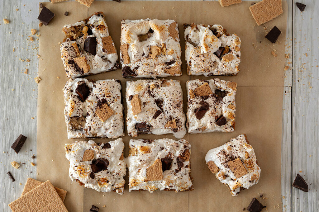 9 S'mores Bars on brown parchment paper. Graham cracker pieces and chocolate pieces are scattered around the bars.