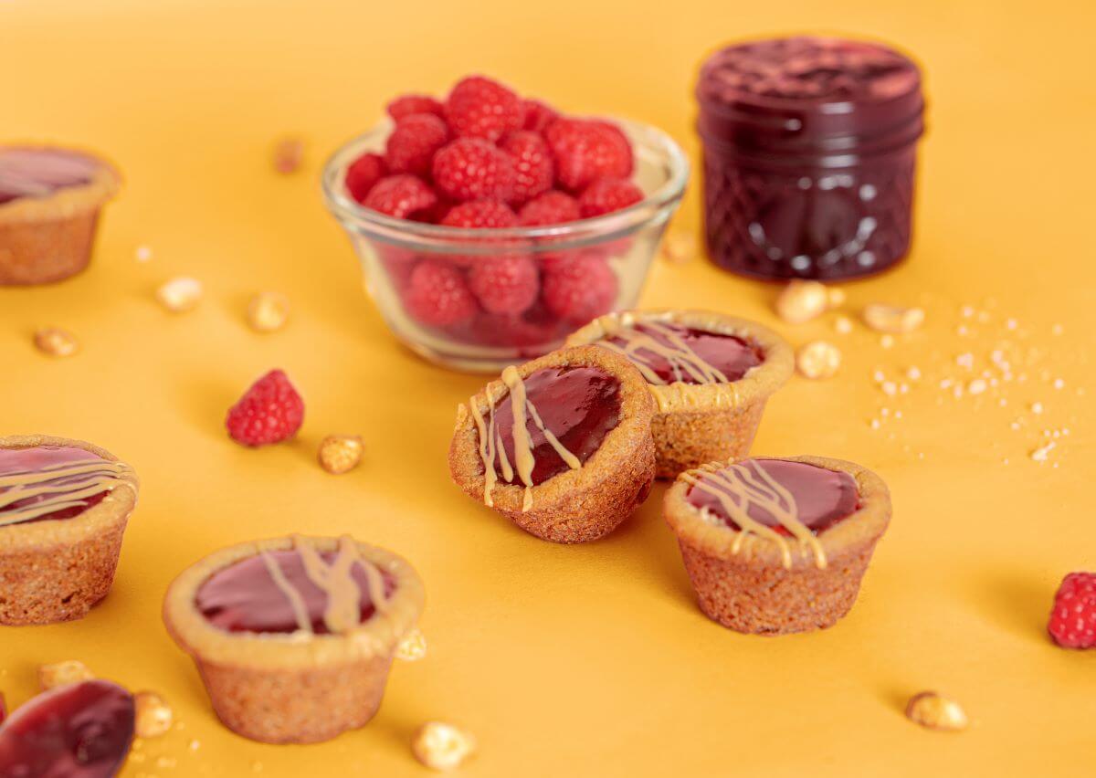 Peanut Butter and Jelly Cookie Cups on orange background. Around the cups are a bowl of raspberries, peanuts, and a jar of raspberry jam.