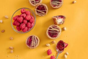 Peanut Butter and Jelly Cookie Cups on orange background. Around the cups are a bowl of raspberries, peanuts, and a spoon with raspberry jam.