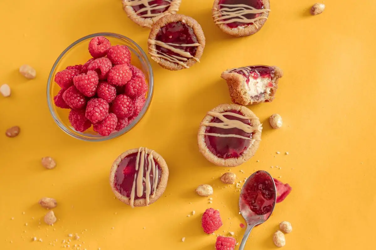 Peanut Butter and Jelly Cookie Cups on orange background. Around the cups are a bowl of raspberries, peanuts, and a spoon with raspberry jam.