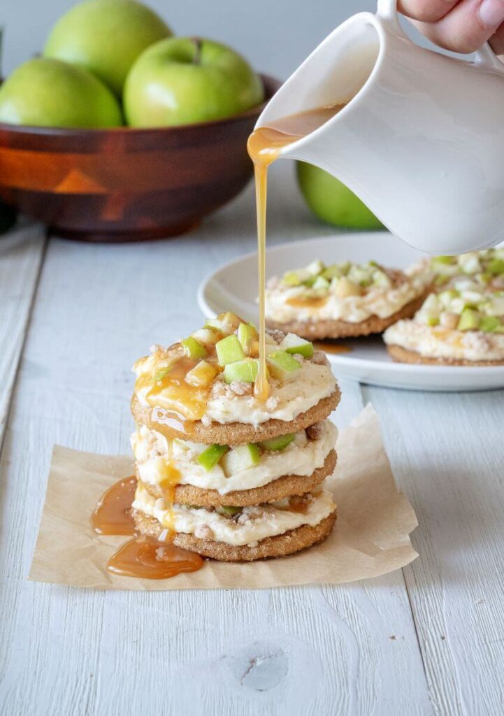 A stack of three Caramel Apple Cookies on a piece of parchment paper. Caramel is being poured on them from above. A plate of cookies and a bowl of green apples are in the background.