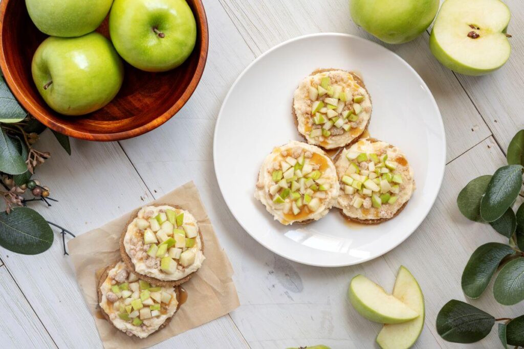 A plate with three Caramel Apple Cookies next to a piece of parchment paper with two cookies on it. There's also a bowl of green apples and slices green applies on the edges of the image.