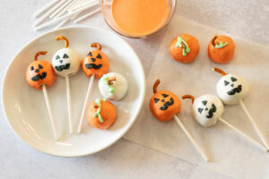 Several Jack O' Lantern Cookie Balls on a plate and several more on parchment paper next to the plate. There's a bowl of sprinkles in the background.