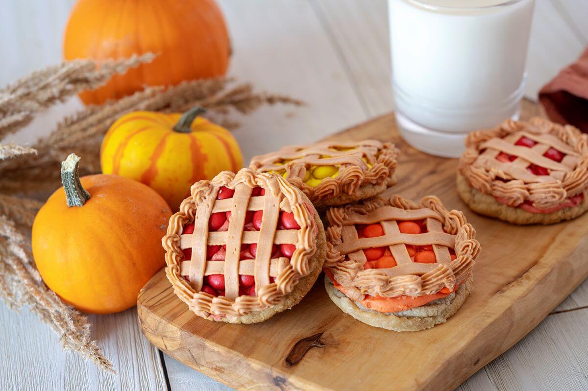 Four Thanksgiving Pie Cookies on a wood serving board with a glass of milk. Several pumpkins and autumn foliage surrounds the board.