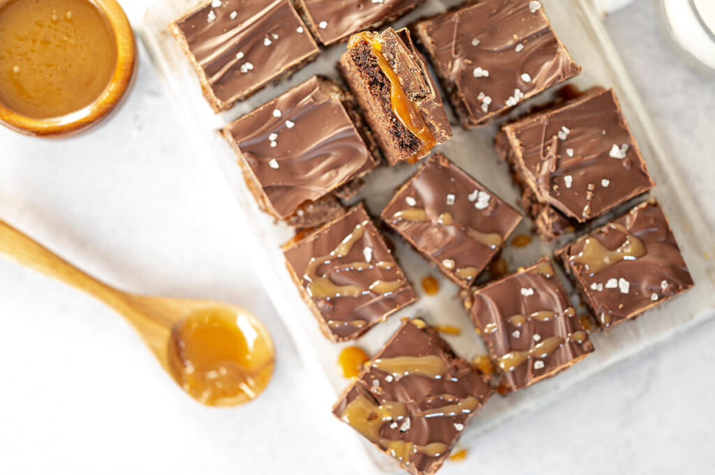 Mocha Caramel Crunch Triple Chocolate Cookies Bars on cutting board next to a bowl of caramel and a wooden spoon.