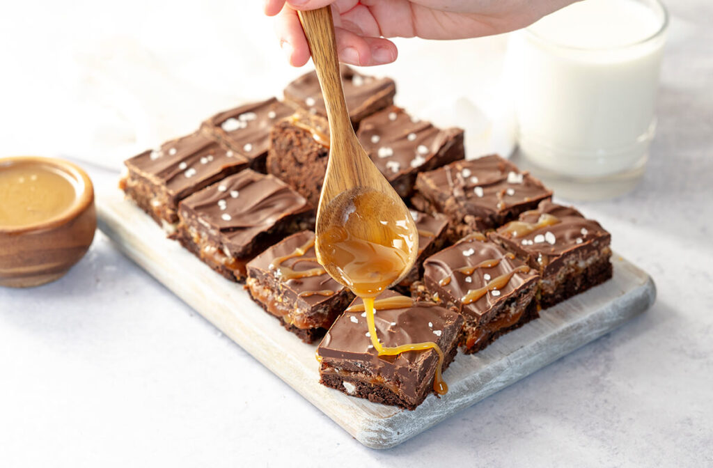 Mocha Caramel Crunch Triple Chocolate Cookie Bars on a wooden serving platter next to a bowl of caramel and glass of milk. Someone is using a wooden spoon to drizzle caramel on the bars.