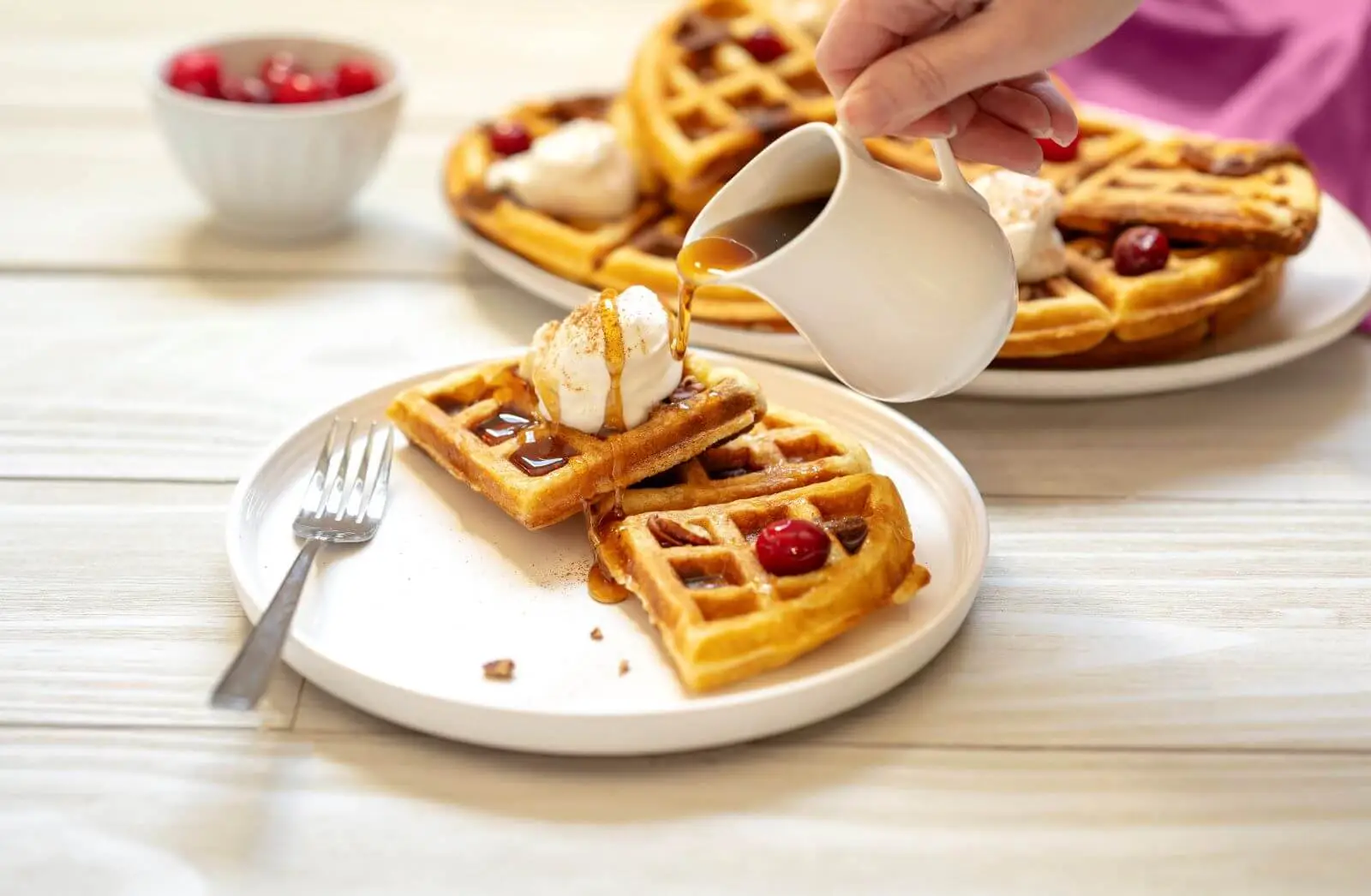 A plate with three Belgian Cookie Dough Waffles topped with homemade whipped cream. Someone is pouring syrup over the waffles. There's a plate full of waffles in the background.