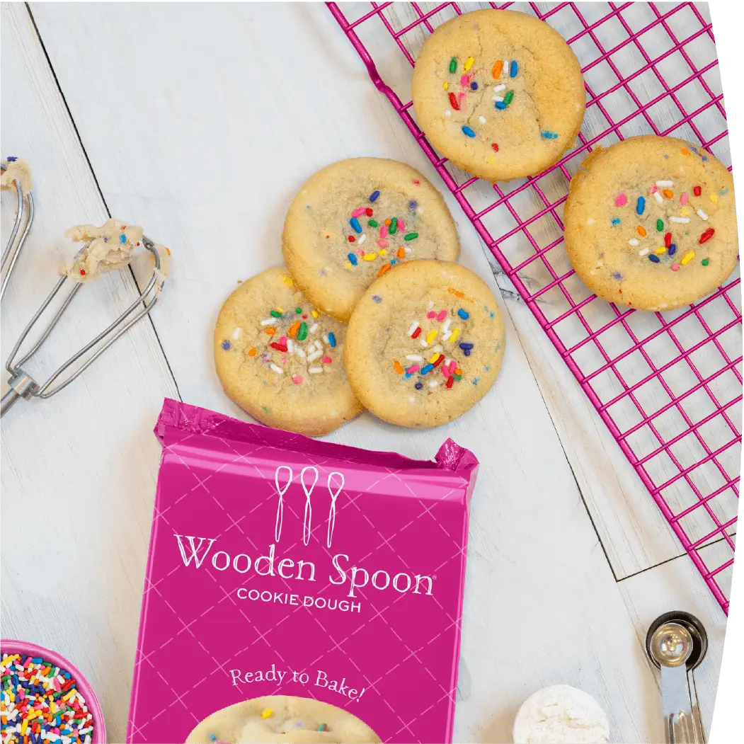 Confetti cookies next to the Wooden Spoon® Cookie Dough Confetti packaging. The cookies are surrounded by cookie dough beaters, measuring spoons, a cooling rack, and a bowl of sprinkles.