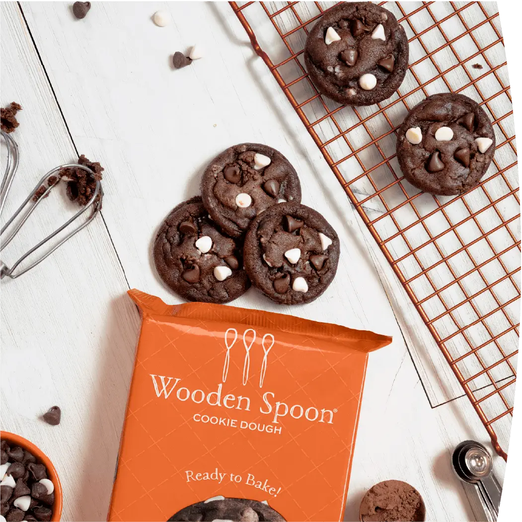 Triple Chocolate cookies next to the Wooden Spoon® Cookie Dough Triple Chocolate packaging. The cookies are surrounded by cookie dough beaters, measuring spoons, a cooling rack, and a bowl of semisweet and white chocolate chips.