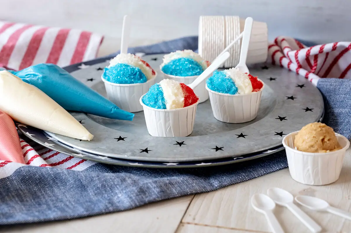 Snow Cone Cookie Balls on a 4th of July themed serving platter next to piping bags filled with icing, an undecorated cookie ball in a cup, and other patriotic decorations.