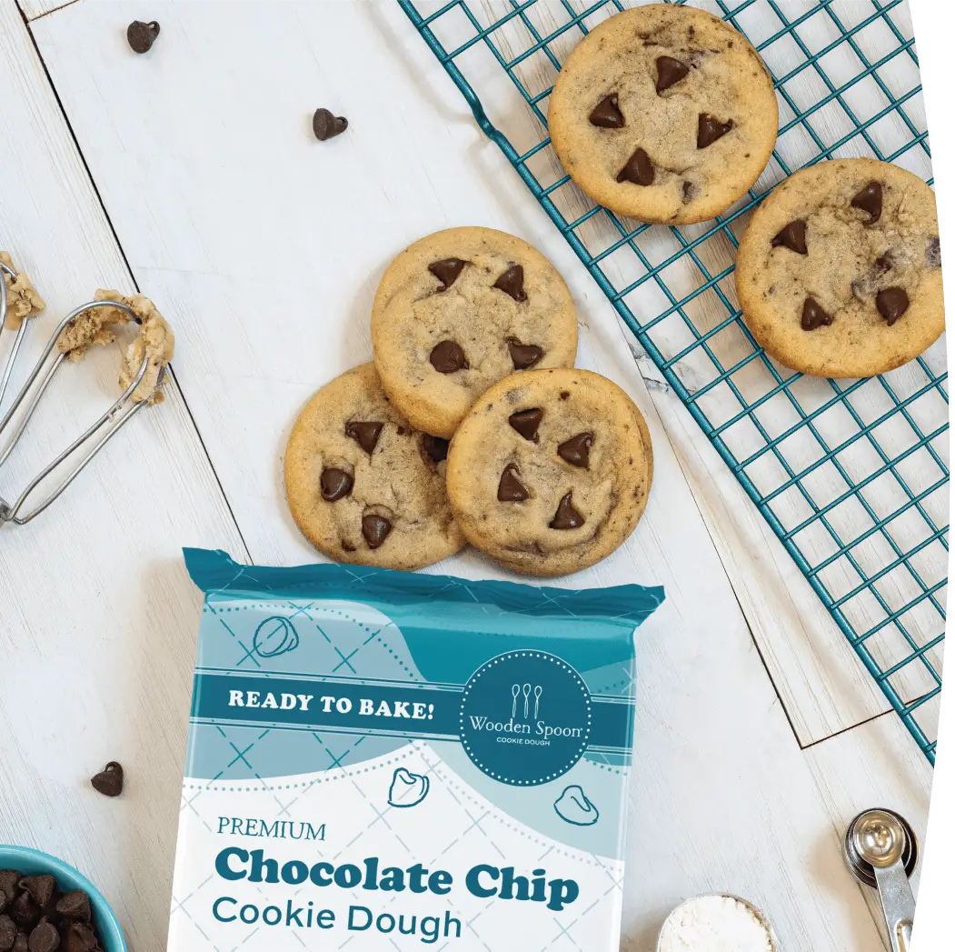 Chocolate Chip cookies next to the Wooden Spoon® Cookie Dough Chocolate Chip packaging. The cookies are surrounded by cookie dough beaters, measuring spoons, a cooling rack, and a bowl of chocolate chips.