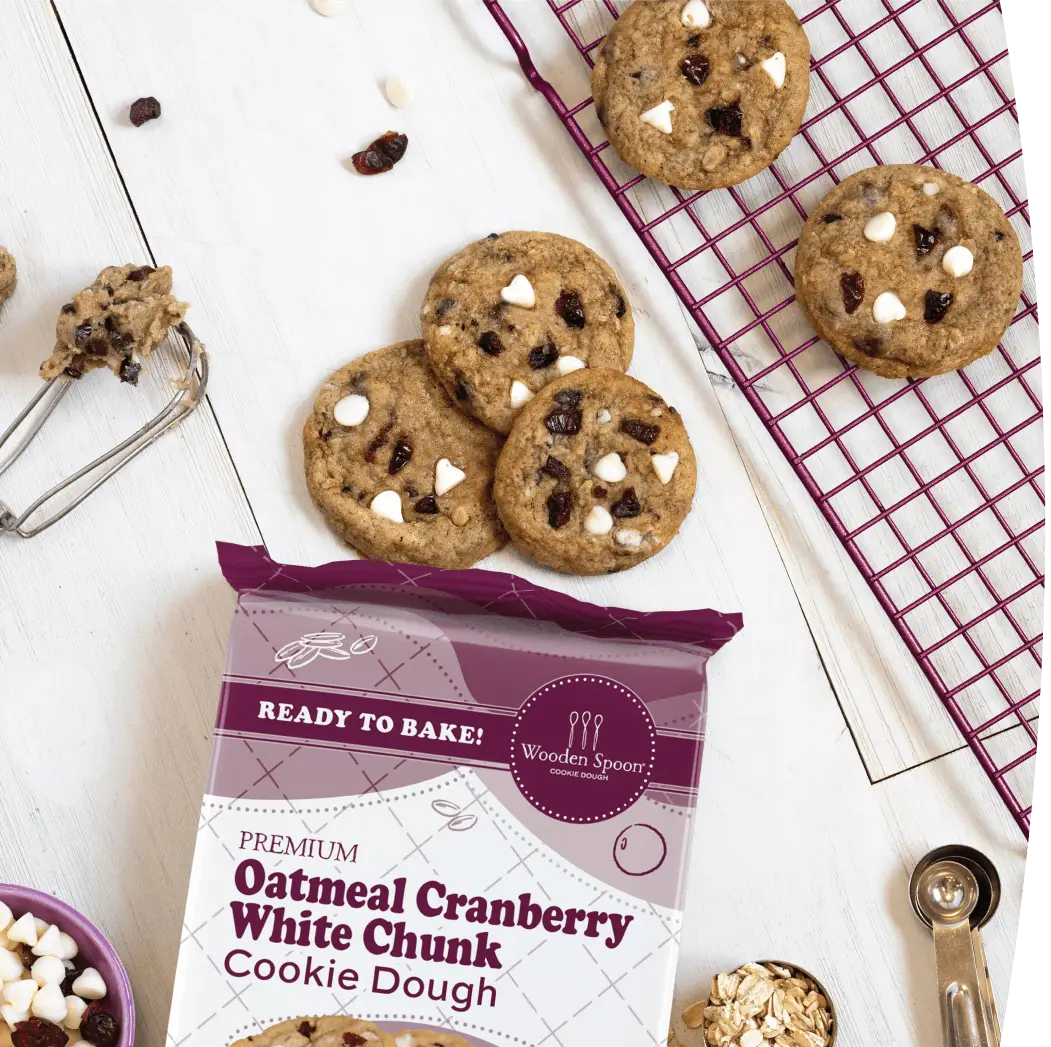 Oatmeal Cranberry cookies next to the Wooden Spoon® Cookie Dough Oatmeal Cranberry packaging. The cookies are surrounded by cookie dough beaters, measuring spoons, a cooling rack, and a bowl of cranberries and white chocolate chips.